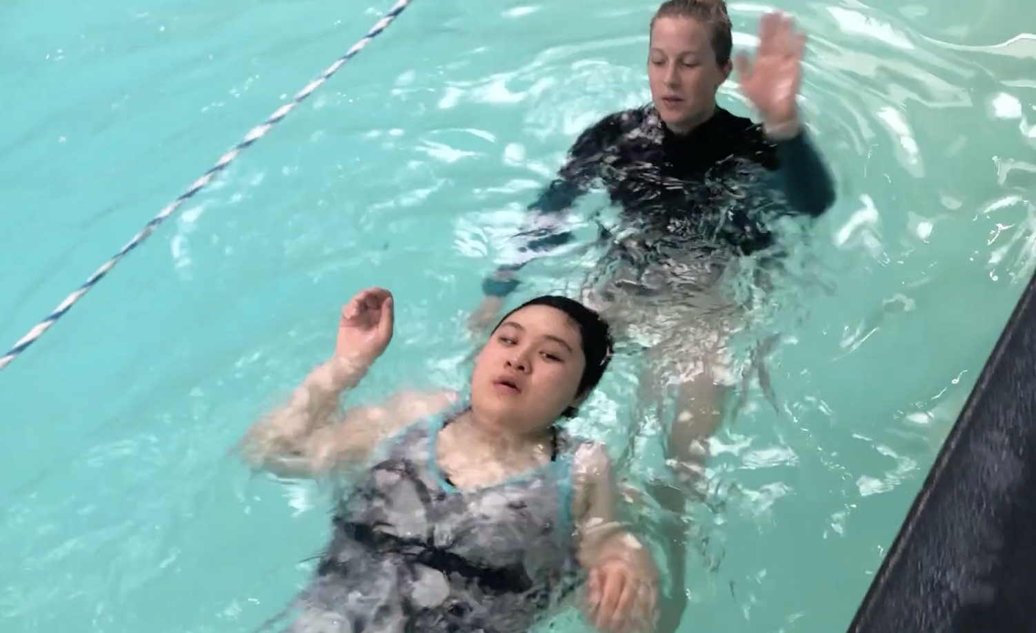 Instructor follows behind a swimmer floating on her back, providing verbal and positional guidance as the swimmer practices backstroke arm movements in the pool.